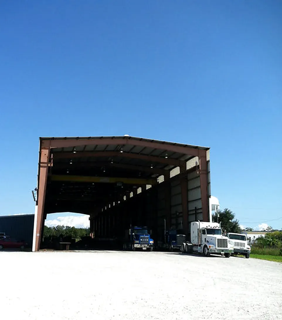 Open-sided fabrication shed with semi trucks and trailers inside, seen from a gravel yard